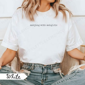 May include: White t-shirt featuring the text "mahjong with mahgirls". The model is wearing blue jeans. The shirt has a crew neck and short sleeves. The word "White" is written in a script font on the bottom left of the image.