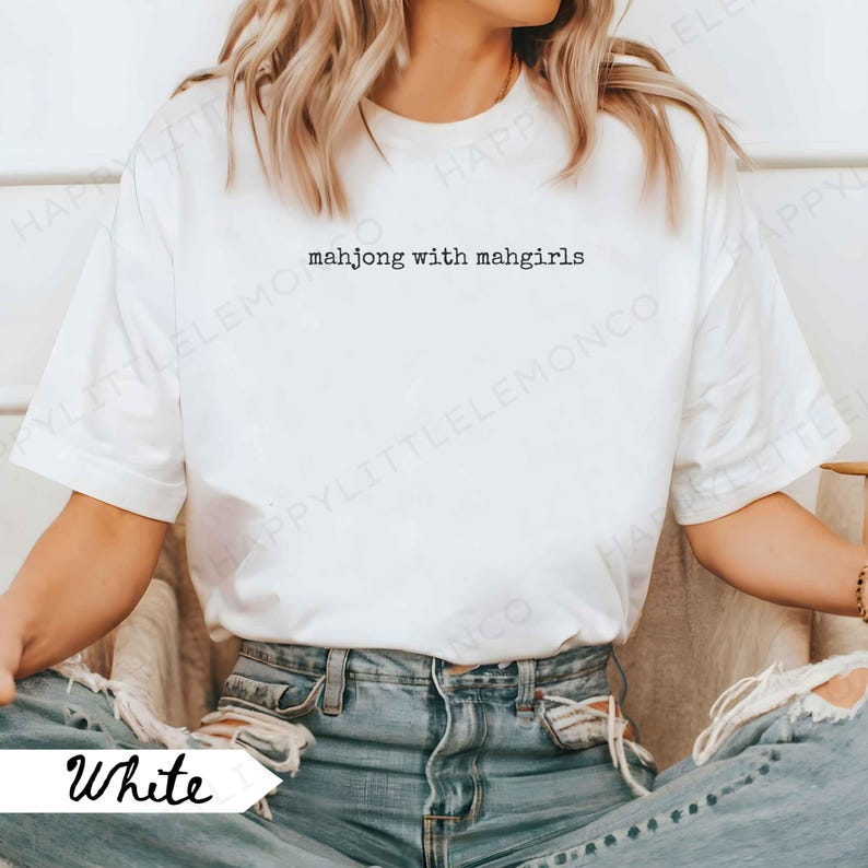 May include: White t-shirt featuring the text "mahjong with mahgirls". The model is wearing blue jeans. The shirt has a crew neck and short sleeves. The word "White" is written in a script font on the bottom left of the image.