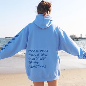 May include: A light blue hoodie with the text "MAKE YOUR HEART THE PRETTIEST THING ABOUT YOU" printed on the back. The left sleeve has a series of blue heart shapes. The hoodie is being worn on a beach.
