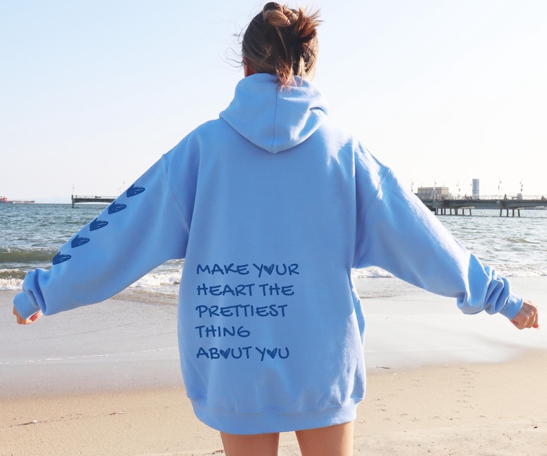 May include: A light blue hoodie with the text "MAKE YOUR HEART THE PRETTIEST THING ABOUT YOU" printed on the back. The left sleeve has a series of blue heart shapes. The hoodie is being worn on a beach.