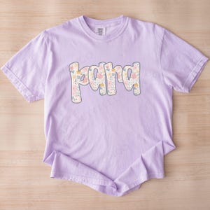 May include: A light purple t-shirt with the word "Mama" in a floral design. The word is in a bubble font with a light blue outline. The flowers are in shades of pink, yellow, and blue.