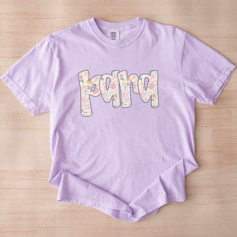 May include: A light purple t-shirt with the word "Mama" in a floral design. The word is in a bubble font with a light blue outline. The flowers are in shades of pink, yellow, and blue.