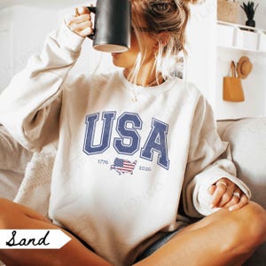 May include: Sand-colored sweatshirt with the word "USA" in navy blue, arched across the chest. Below the text is a small American flag graphic with the years 1776 and 2026. The person is holding a black mug.