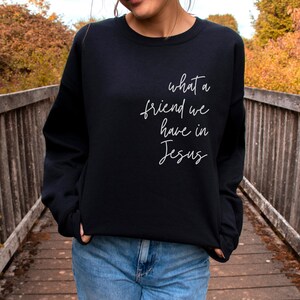 May include: A black long-sleeve crewneck sweatshirt featuring white cursive text on the front that reads, what a friend we have in Jesus. The garment has a ribbed neckline, cuffs, and hem, and is paired with blue denim jeans.