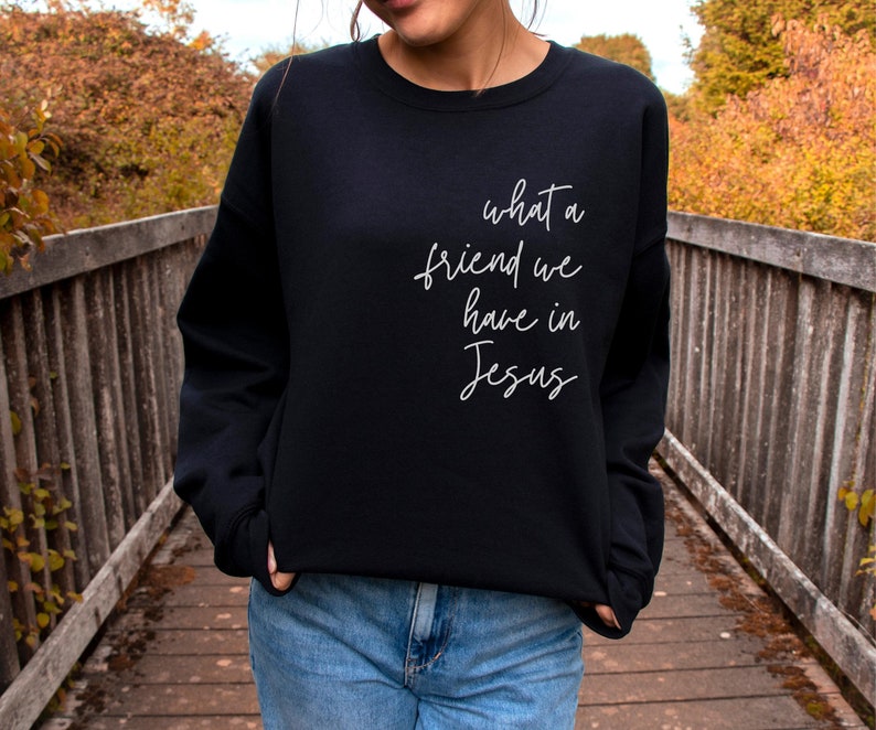 May include: A black long-sleeve crewneck sweatshirt featuring white cursive text on the front that reads, what a friend we have in Jesus. The garment has a ribbed neckline, cuffs, and hem, and is paired with blue denim jeans.