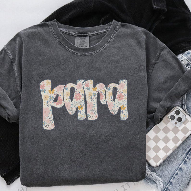 May include: A dark gray, long-sleeved shirt with the word "Mama" in a floral pattern across the front. The shirt is made of a soft material and has a crew neck. A phone with a checkered case and a black jacket are in the background.