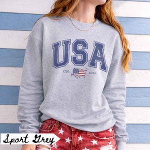 May include: A light gray sweatshirt with the word "USA" in navy blue block letters. Below the word "USA" are the numbers "1776" and "2026", with a small American flag graphic in between. The words "Sport Grey" are at the bottom.