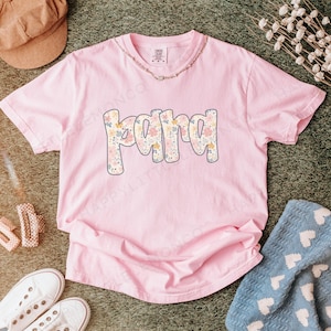 May include: A light pink t-shirt with the word "PAPA" in a floral design. The letters are outlined in a light blue color. The shirt is made of a soft material and is likely a comfort color tee. A gold necklace is visible.
