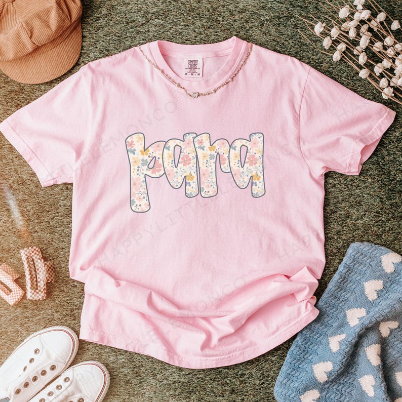 May include: A light pink t-shirt with the word "PAPA" in a floral design. The letters are outlined in a light blue color. The shirt is made of a soft material and is likely a comfort color tee. A gold necklace is visible.