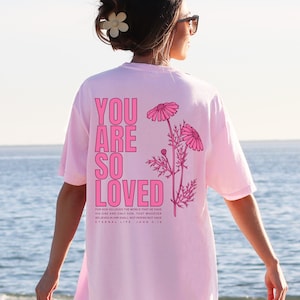 May include: A light pink t-shirt with the words "YOU ARE SO LOVED" in bright pink block letters. The shirt also features a floral design and a Bible verse. The shirt is worn by a person with sunglasses and a flower in their hair.