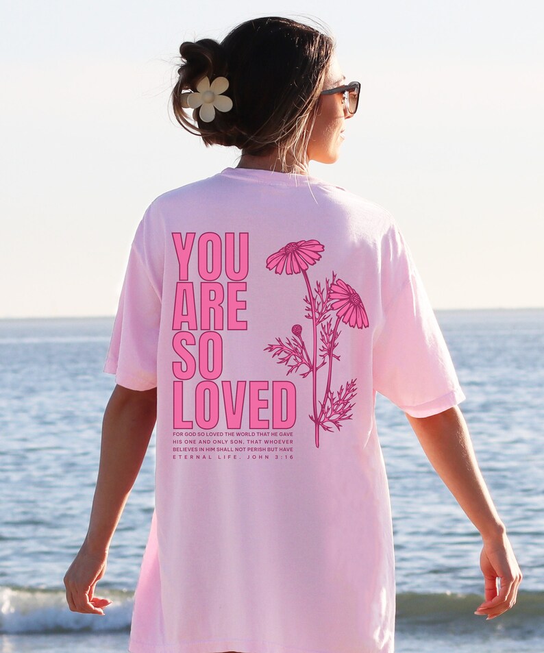 May include: A light pink t-shirt with the words "YOU ARE SO LOVED" in bright pink block letters. The shirt also features a floral design and a Bible verse. The shirt is worn by a person with sunglasses and a flower in their hair.