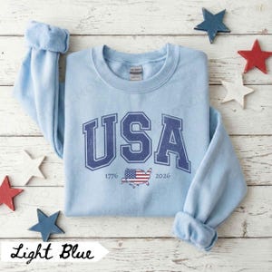 May include: Light blue sweatshirt with the word "USA" in navy blue, along with the years "1776" and "2026" and an American flag graphic. The sweatshirt is surrounded by wooden star decorations in red, white, and blue.
