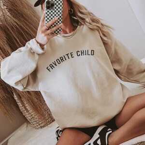 May include: A beige crewneck sweatshirt featuring the text "FAVORITE CHILD" in black, arched lettering across the chest. The garment has long sleeves with ribbed cuffs and a ribbed neckline, constructed from a solid-colored fabric.