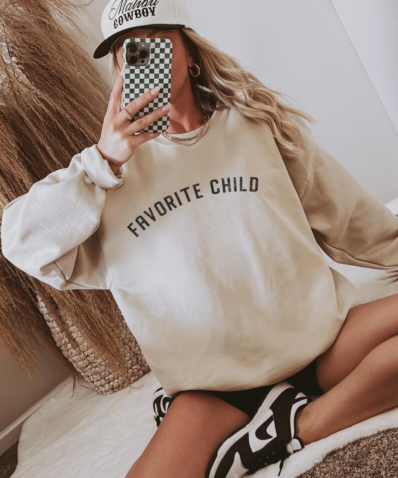 May include: A beige crewneck sweatshirt featuring the text "FAVORITE CHILD" in black, arched lettering across the chest. The garment has long sleeves with ribbed cuffs and a ribbed neckline, constructed from a solid-colored fabric.