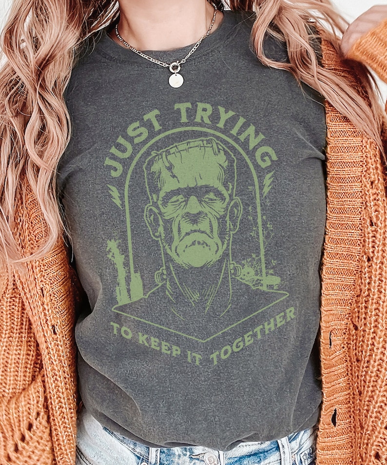 May include: A dark grey t-shirt with a green graphic of Frankenstein's monster. The text "Just Trying To Keep It Together" is printed in green around the monster's head.