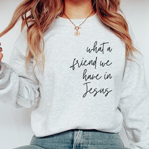 May include: A light grey crewneck sweatshirt featuring the black cursive text what a friend we have in Jesus printed on the front. The garment has long sleeves and a ribbed neckline, cuffs, and hem, providing a simple and clean aesthetic.