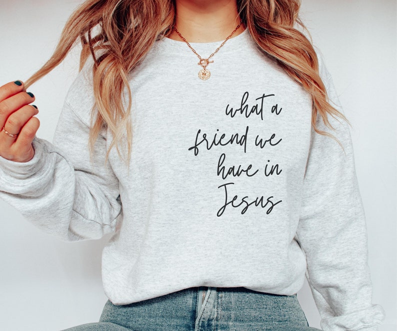 May include: A light grey crewneck sweatshirt featuring the black cursive text what a friend we have in Jesus printed on the front. The garment has long sleeves and a ribbed neckline, cuffs, and hem, providing a simple and clean aesthetic.