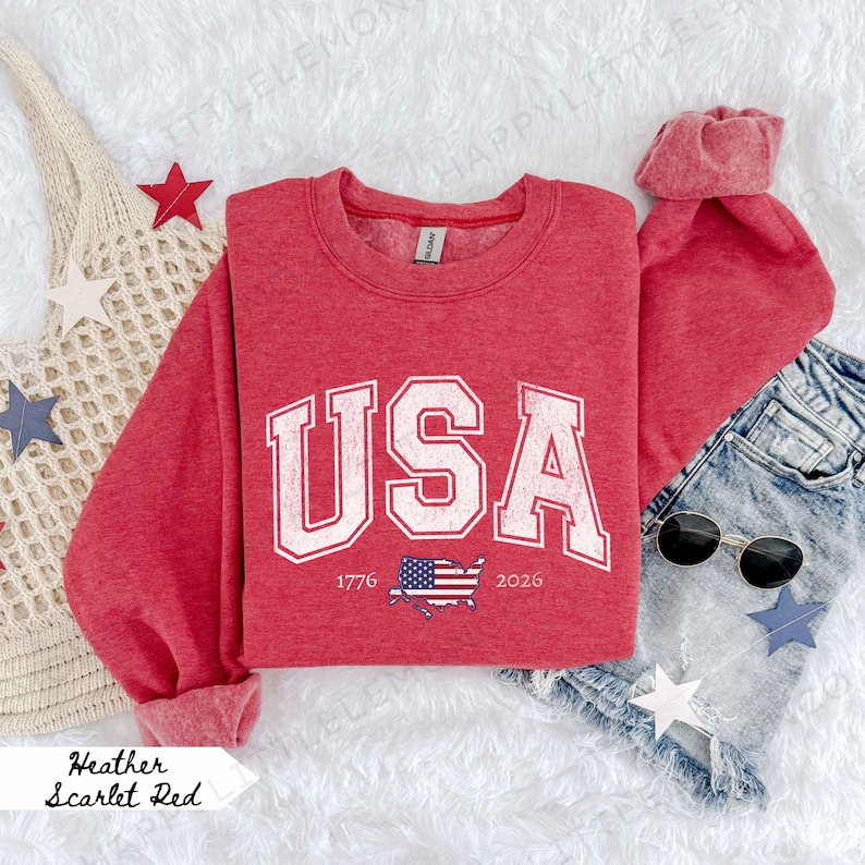 May include: A red heather sweatshirt with the word "USA" in a white, collegiate-style font. Below the text is a graphic of the American flag and the years 1776 and 2026. The sweatshirt is paired with denim shorts and sunglasses.
