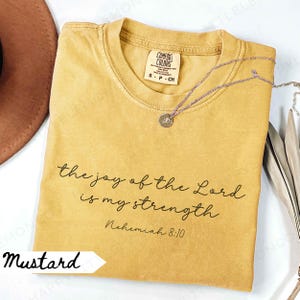 May include: Mustard-colored t-shirt with the text "the joy of the Lord is my strength" and "Nehemiah 8:10". A silver necklace with a small pendant rests on the shirt. The shirt is labeled "Comfort Colors".