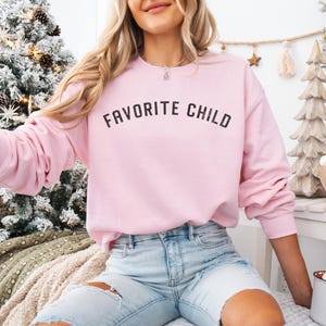 May include: A light pink crewneck sweatshirt featuring the text FAVORITE CHILD printed in a distressed black font across the chest. The long-sleeved garment has a ribbed neckline, cuffs, and hem, providing a classic fit for casual wear.