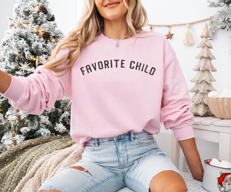 May include: A light pink crewneck sweatshirt featuring the text FAVORITE CHILD printed in a distressed black font across the chest. The long-sleeved garment has a ribbed neckline, cuffs, and hem, providing a classic fit for casual wear.