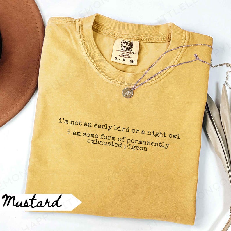 May include: Mustard-colored Comfort Colors t-shirt with the text "i'm not an early bird or a night owl i am some form of permanently exhausted pigeon." A silver necklace with a pendant is on the shirt.