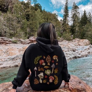 May include: A black hooded sweatshirt with a colorful mushroom and frog design on the back. The design features various types of mushrooms, a frog, a snail, leaves, and pine cones.
