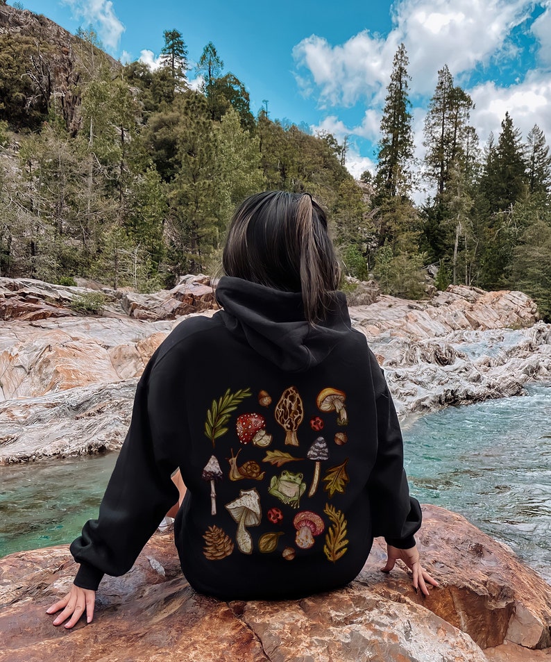 May include: A black hooded sweatshirt with a colorful mushroom and frog design on the back. The design features various types of mushrooms, a frog, a snail, leaves, and pine cones.