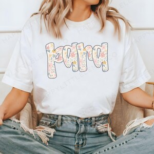 May include: White t-shirt with the word "Mama" in a floral design. The word is in a bubble font with a light blue outline. The floral design includes pink, yellow, and blue flowers. The model is wearing blue jeans.