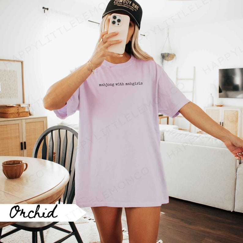 May include: A lilac t-shirt with the text "mahjong with mahgirls" in black. The shirt is a light purple colour. A black baseball cap is worn. The word "Orchid" is written in a script font at the bottom.