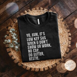 May include: Black t-shirt with white text: "YO, FAM, IT'S LOW KEY SUS WHEN U DON'T SHOW UR WORK. NO CAP. DO BETTER, BESTIE." The shirt is folded on a woven, circular placemat with a wooden background. Eucalyptus sprigs are on the left.