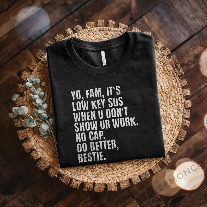 May include: Black t-shirt with white text: "YO, FAM, IT'S LOW KEY SUS WHEN U DON'T SHOW UR WORK. NO CAP. DO BETTER, BESTIE." The shirt is folded on a woven, circular placemat with a wooden background. Eucalyptus sprigs are on the left.