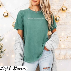 May include: Light green t-shirt with the text "mahjong with mahgirls" in white. The shirt is a solid colour and has short sleeves. A woman is wearing the shirt with a grey cardigan draped over her shoulders.