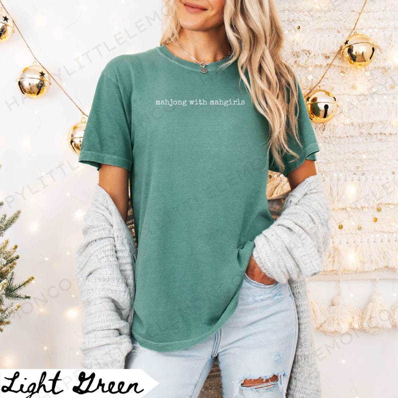 May include: Light green t-shirt with the text "mahjong with mahgirls" in white. The shirt is a solid colour and has short sleeves. A woman is wearing the shirt with a grey cardigan draped over her shoulders.