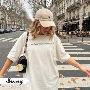 May include: Ivory-colored t-shirt with the text "mahjong with mahsisters" in black. A beige baseball cap with a black logo is worn. A black purse with a gold chain is also visible.