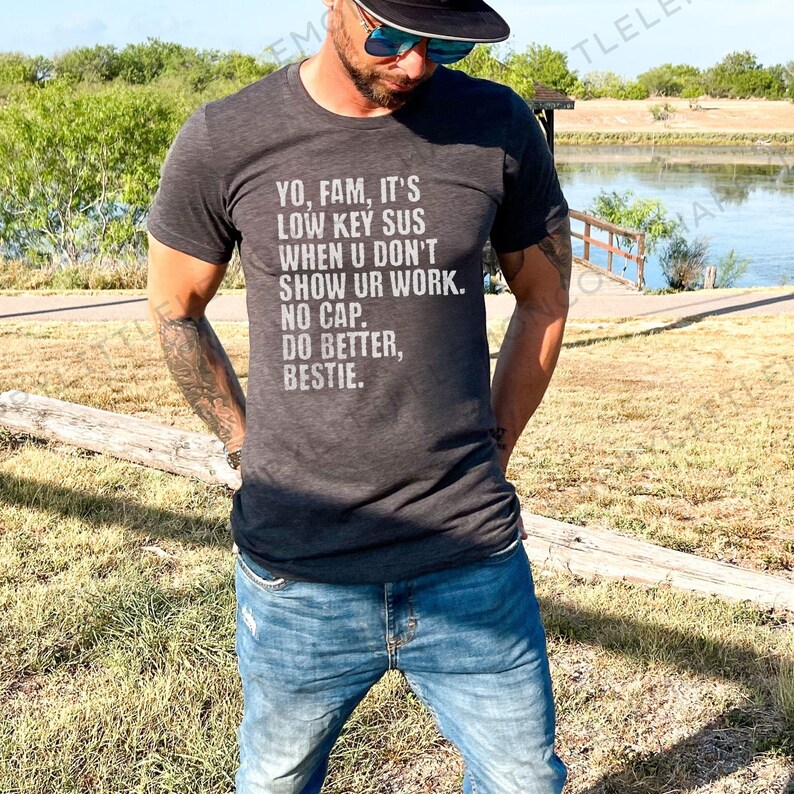 May include: Dark gray t-shirt with the text "YO, FAM, IT'S LOW KEY SUS WHEN U DON'T SHOW UR WORK. NO CAP. DO BETTER, BESTIE." The shirt is paired with blue jeans. The person is wearing sunglasses and a black hat.