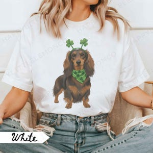 May include: White t-shirt featuring a dachshund wearing a green bandana and a shamrock headband. The dog is brown and black. The word "White" is printed on a white banner in the lower left corner.