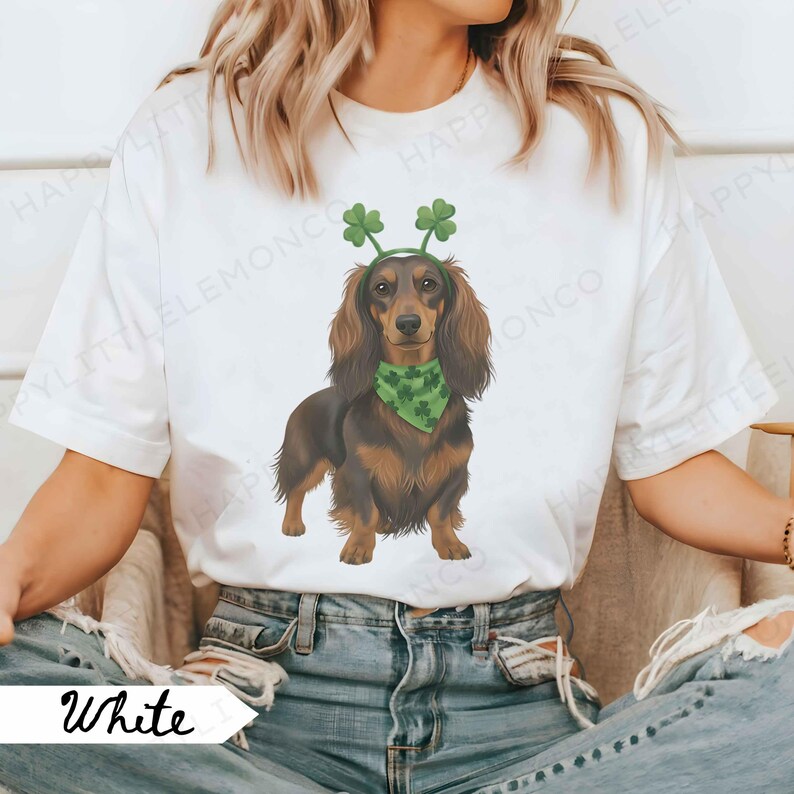 May include: White t-shirt featuring a dachshund wearing a green bandana and a shamrock headband. The dog is brown and black. The word "White" is printed on a white banner in the lower left corner.