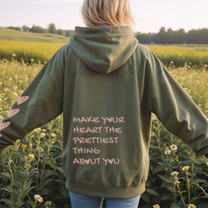 May include: Olive green hoodie with the text "MAKE YOUR HEART THE PRETTIEST THING ABOUT YOU" in pink. The hoodie has a hood and a pink heart design on the left sleeve. The person is standing in a field of flowers.