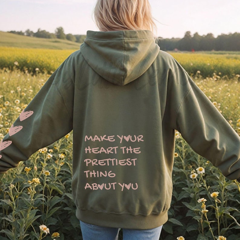 May include: Olive green hoodie with the text "MAKE YOUR HEART THE PRETTIEST THING ABOUT YOU" in pink. The hoodie has a hood and a pink heart design on the left sleeve. The person is standing in a field of flowers.
