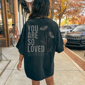 May include: Black oversized t-shirt with the text "YOU ARE SO LOVED" in large, gray letters. Below the text are the words "FOR GOD SO LOVED THE WORLD... ETERNAL LIFE. JOHN 3:16". The shirt also features a floral design.