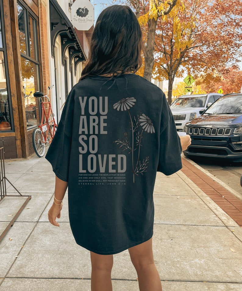 May include: Black oversized t-shirt with the text "YOU ARE SO LOVED" in large, gray letters. Below the text are the words "FOR GOD SO LOVED THE WORLD... ETERNAL LIFE. JOHN 3:16". The shirt also features a floral design.
