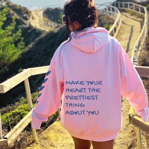 May include: A light pink hoodie with the text "MAKE YOUR HEART THE PRETTIEST THING ABOUT YOU" in blue. The hoodie has a hood and long sleeves with blue arrow designs on the left sleeve.