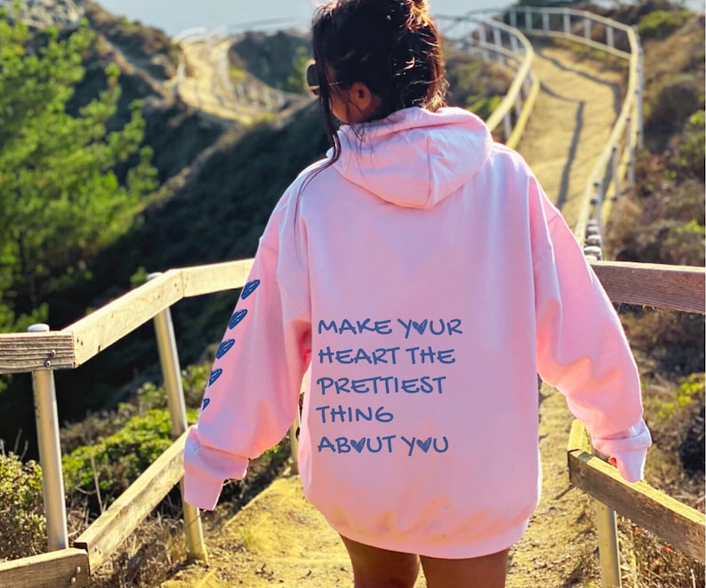 May include: A light pink hoodie with the text "MAKE YOUR HEART THE PRETTIEST THING ABOUT YOU" in blue. The hoodie has a hood and long sleeves with blue arrow designs on the left sleeve.