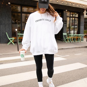 May include: A white crewneck sweatshirt with the text "FAVORITE CHILD" printed in black across the chest, paired with black leggings, white socks, white sneakers, and a black baseball cap featuring the embroidered white text "11:11".