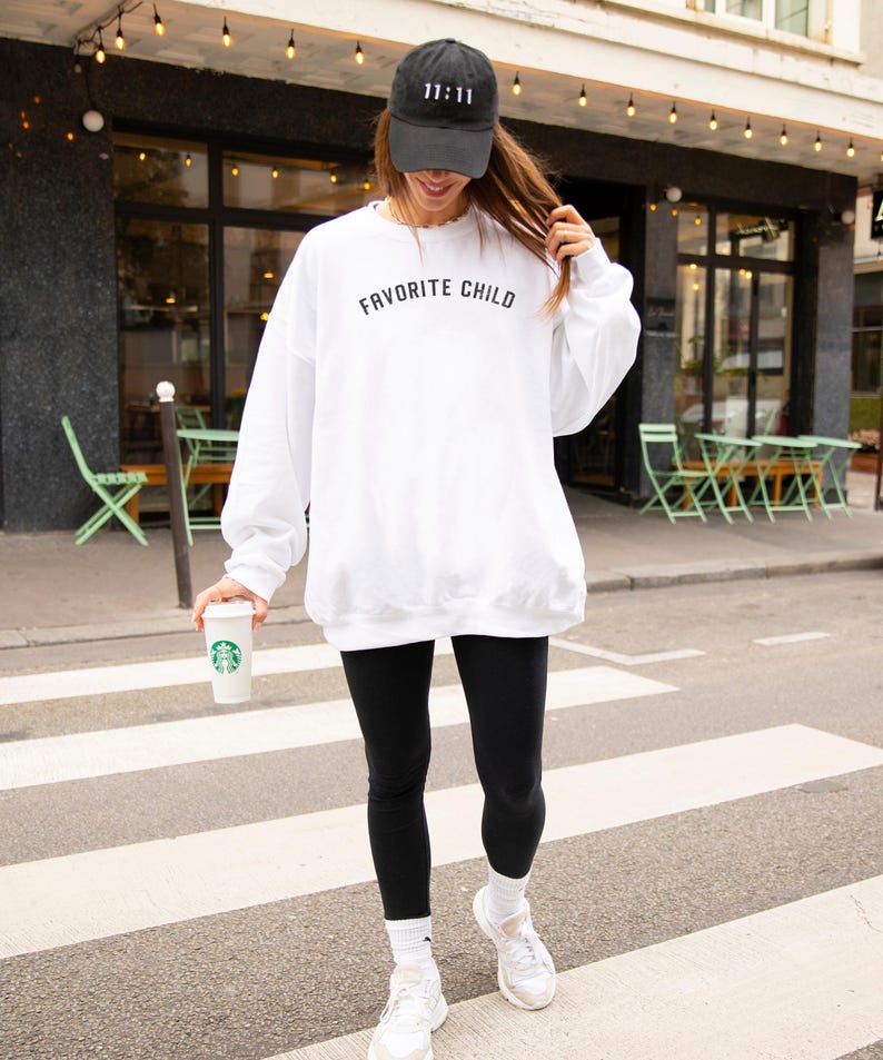 May include: A white crewneck sweatshirt with the text "FAVORITE CHILD" printed in black across the chest, paired with black leggings, white socks, white sneakers, and a black baseball cap featuring the embroidered white text "11:11".