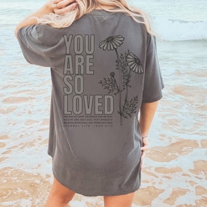 May include: Dark gray t-shirt with the words "YOU ARE SO LOVED" printed on the back in a bold, block font. Below the text is a floral illustration and a Bible verse. The shirt is a casual style.
