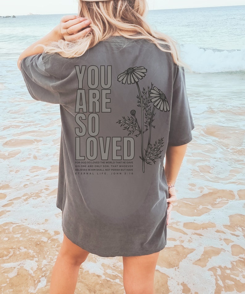May include: Dark gray t-shirt with the words "YOU ARE SO LOVED" printed on the back in a bold, block font. Below the text is a floral illustration and a Bible verse. The shirt is a casual style.