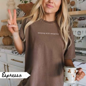 May include: Espresso-coloured t-shirt with the text "mahjong with mahgirls" in white. The shirt is a solid colour and has a crew neck. A white mug with brown bear graphics is held in the hand.