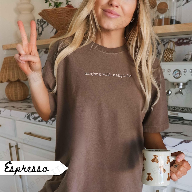 May include: Espresso-coloured t-shirt with the text "mahjong with mahgirls" in white. The shirt is a solid colour and has a crew neck. A white mug with brown bear graphics is held in the hand.